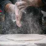 A chef clapping hands over dough, releasing flour in a kitchen setting.